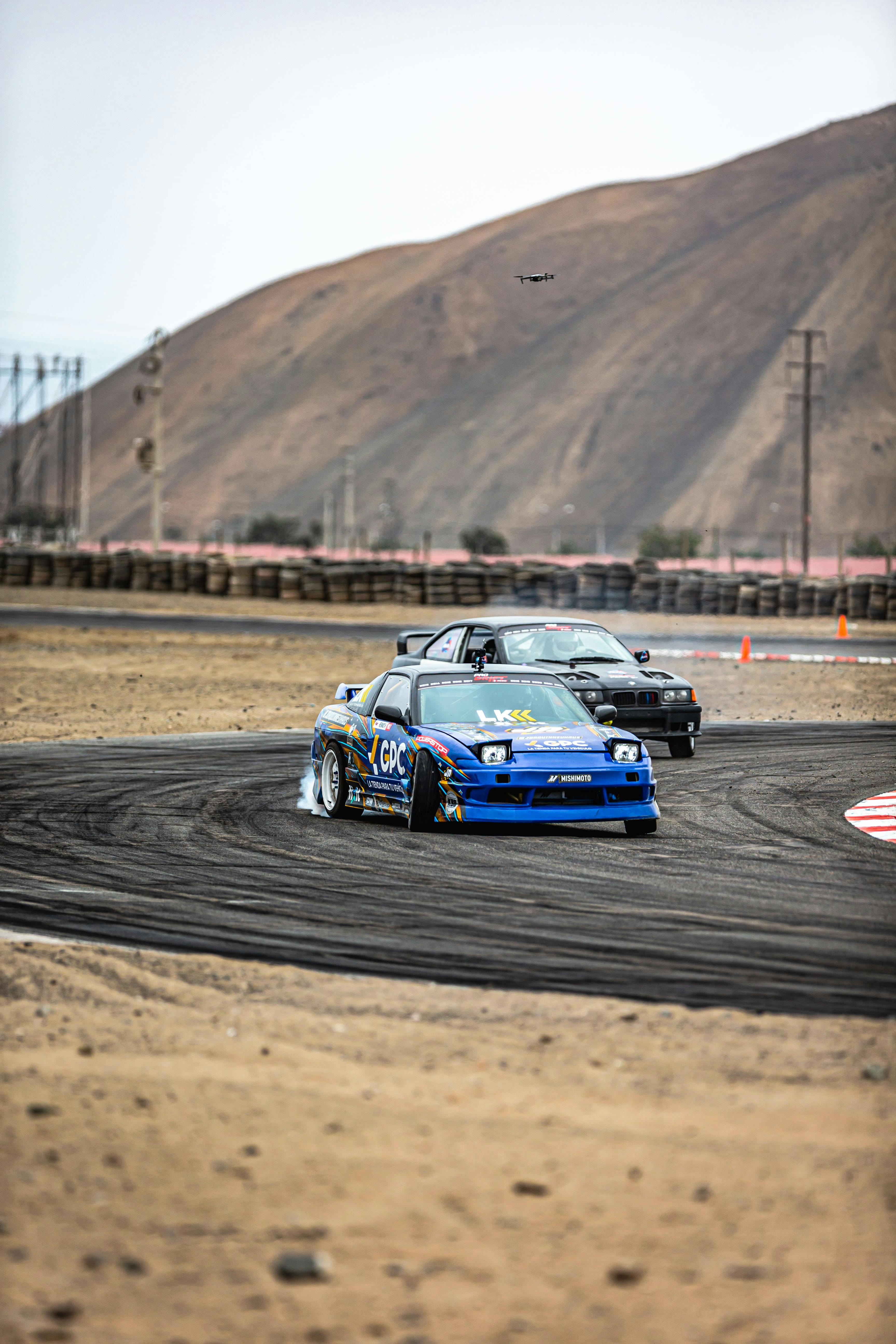 Two cars drifting on a track with mountains in the background.